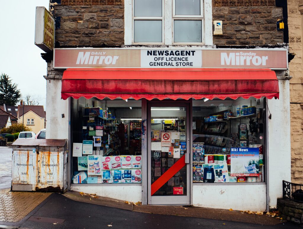 The picture shows a grotty shop front with a dirty, red awning. The windows are full of posters and signs. To the left of the shop are two filthy bins with rust staining..